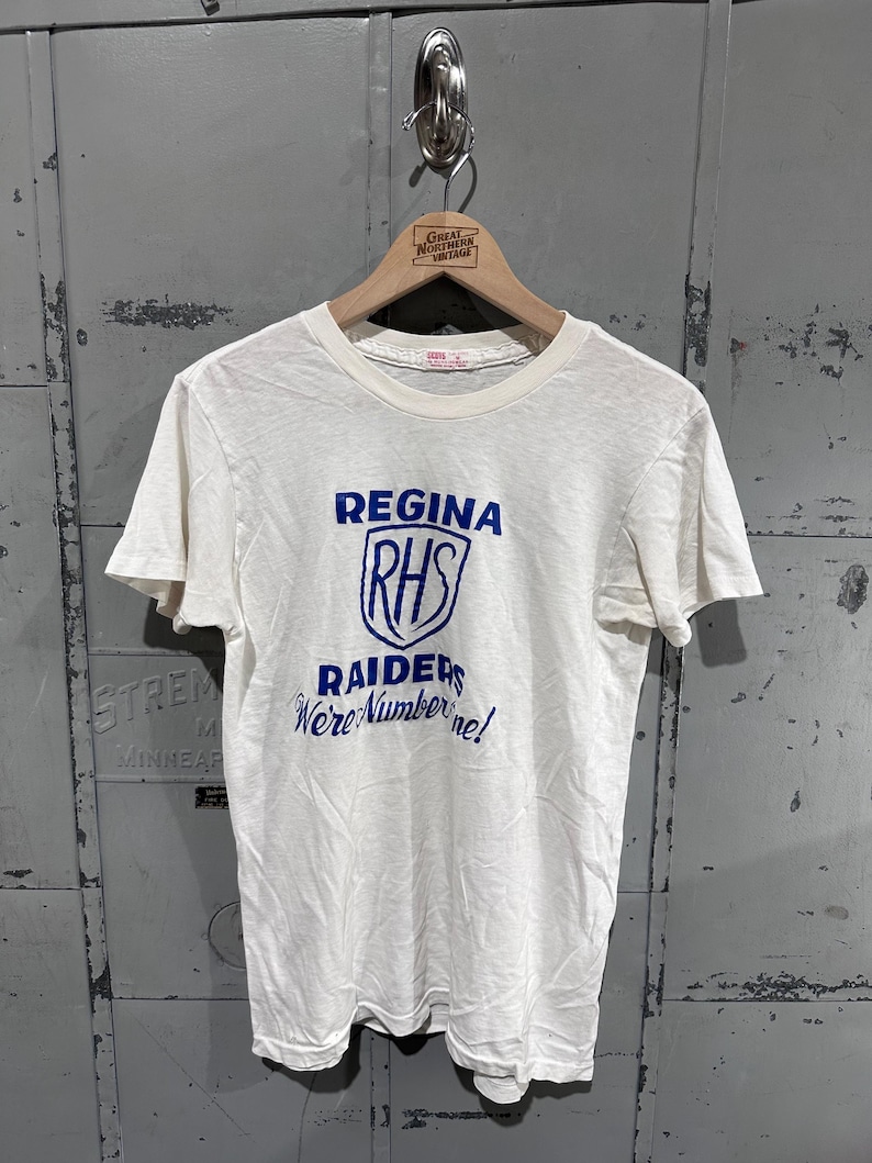 May include: Cream-colored t-shirt with the text "Regina RHS Raiders We're Number One!" in blue. The design features a shield graphic. The shirt is hanging on a wooden hanger.