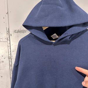 May include: A navy blue hooded sweatshirt with a large hood. The sweatshirt has a drawstring and a kangaroo pocket. The fabric appears to be a thick, durable material. There are small holes in the fabric.