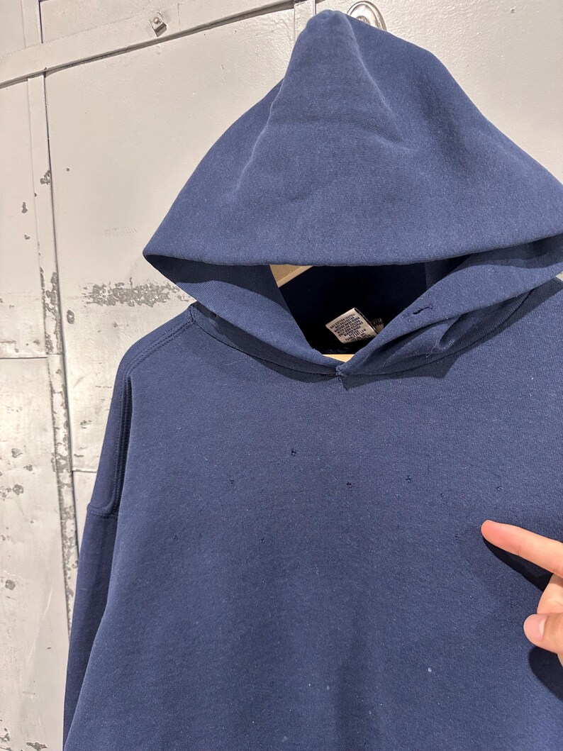 May include: A navy blue hooded sweatshirt with a large hood. The sweatshirt has a drawstring and a kangaroo pocket. The fabric appears to be a thick, durable material. There are small holes in the fabric.