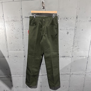 Puede incluir: Pantalones verde oliva con cierre de botón, bolsillos delanteros y trabillas para el cinturón. Los pantalones cuelgan de una percha de madera sobre un fondo metálico texturizado. Los pantalones parecen estar hechos de una tela duradera.