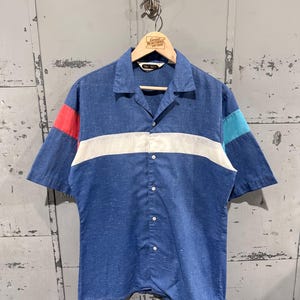 May include: Blue short-sleeve button-down shirt with a white horizontal stripe across the chest. The sleeves have red and teal color blocks. The shirt is hanging on a wooden hanger with the text "Great Northern Vintage".