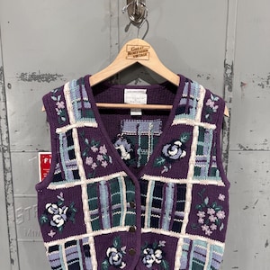 May include: A vintage purple knit vest with a grid pattern, featuring embroidered floral designs in white, blue, and pink. The vest has a V-neck, button closure, and is displayed on a wooden hanger. The label reads "Great Northern Vintage".