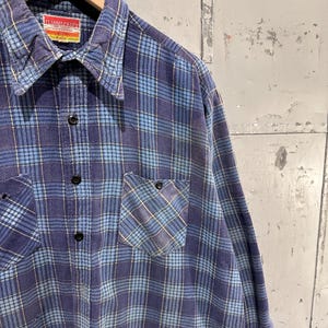 Pu&ograve; includere: Una camicia a maniche lunghe abbottonata con un motivo a quadri blu e azzurro. La camicia presenta un colletto a punta, due tasche sul petto con chiusura a bottone e un'etichetta rossa e bianca sul collo con la scritta Flannelcraft.