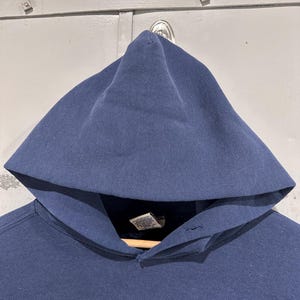 May include: A navy blue hooded sweatshirt with a large hood. The sweatshirt is hanging on a wooden hanger. The fabric appears to be a soft, medium-weight material. There are small holes in the fabric.