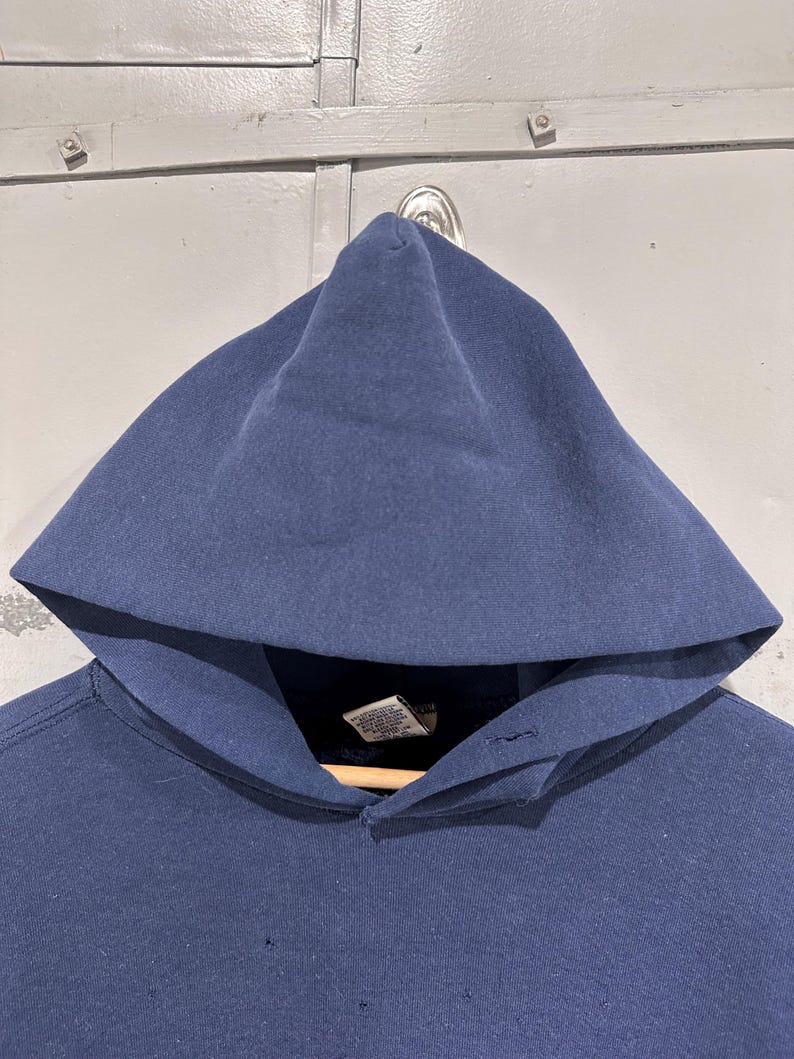 May include: A navy blue hooded sweatshirt with a large hood. The sweatshirt is hanging on a wooden hanger. The fabric appears to be a soft, medium-weight material. There are small holes in the fabric.