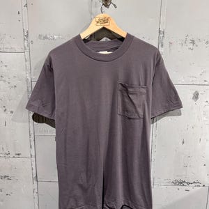 May include: A solid dark charcoal gray short-sleeve t-shirt featuring a crew neckline and a single patch pocket on the left side of the chest. The garment is displayed on a wooden hanger against a gray metal wall with visible panel seams.