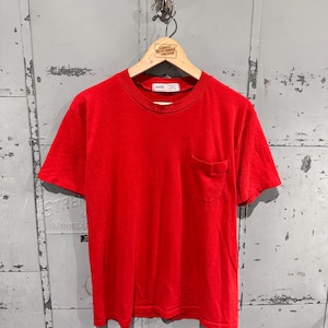 May include: A plain red t-shirt with a chest pocket, displayed on a wooden hanger. The short-sleeved shirt is made of a soft fabric. The hanger is suspended from a metal hook on a distressed metal surface.