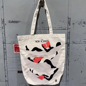May include: Cream-colored tote bag featuring the text "THE NEW YORKER" above an illustration of figures reading books. The illustration has black figures with red books and light gray wavy lines. The bag has two cream-colored handles.