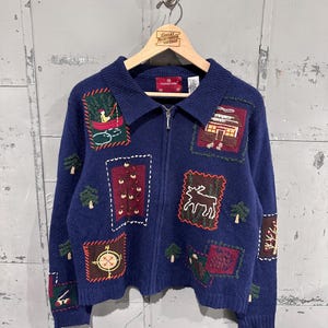 Op de afbeelding: Een marineblauw gebreid vest met rits, een kraag en diverse geborduurde patches met buitenmotieven, waaronder een persoon in een rode kano, een blokhut, een eland, een kompas en dennenbomen, met het Holiday Lodge label aan de binnenzijde.