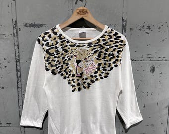 May include: White three-quarter sleeve top with a leopard print design across the chest. The leopard print features black, tan, and gold spots, with a detailed leopard face in the center. The top is hanging on a wooden hanger.