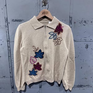 May include: A tan zip-up sweater with a collared neckline. The sweater features embroidered leaf designs in shades of blue, purple, and pink. The sweater is hanging on a wooden hanger with the text "Great Northern Vintage".