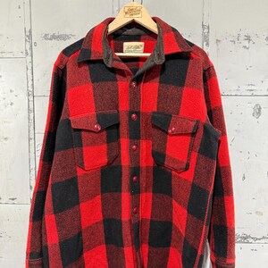 May include: A red and black plaid flannel shirt with two chest pockets and red buttons. The shirt is hanging on a wooden hanger with a tag that reads "Great Northern Vintage".