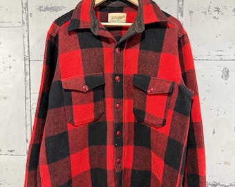 May include: A red and black plaid flannel shirt with two chest pockets and red buttons. The shirt is hanging on a wooden hanger with a tag that reads "Great Northern Vintage".