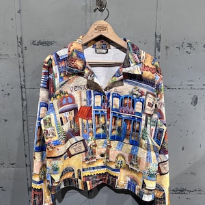 Peut inclure: Une chemise boutonnée avec un imprimé coloré représentant des scènes de rue vénitiennes. La chemise a un col, des manches longues et une fermeture boutonnée. L'imprimé comprend des bâtiments, des boutiques et le mot "Venice".