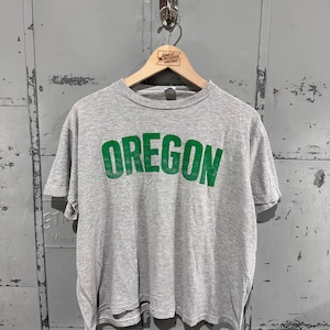 Puede incluir: Una camiseta gris jaspeado de manga corta con la palabra OREGON impresa en una fuente verde desgastada en el pecho. La prenda tiene cuello redondo y está colgada en una percha de madera con el texto Great Northern Vintage.