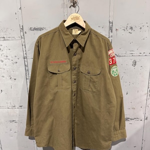 Pode incluir: Camisa verde-oliva de manga comprida com dois bolsos no peito e botões. Apresenta texto vermelho no bolso esquerdo e um patch com o número 37 e um emblema verde na manga direita. A camisa está pendurada num cabide de madeira.