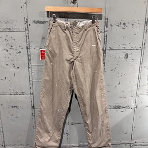 May include: Khaki-colored pants with white paint splatters hanging on a wooden hanger. The pants have a button and zipper closure, belt loops, and pockets. The pants are made of a durable fabric and appear to be vintage or workwear style.