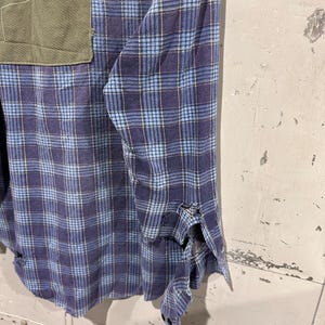 Pu&ograve; includere: Una camicia a maniche lunghe con bottoni e motivo a quadri blu e viola. La camicia presenta una tasca applicata rettangolare color verde oliva sul petto e mostra visibili sfilacciature e danni al tessuto lungo il polsino della manica.