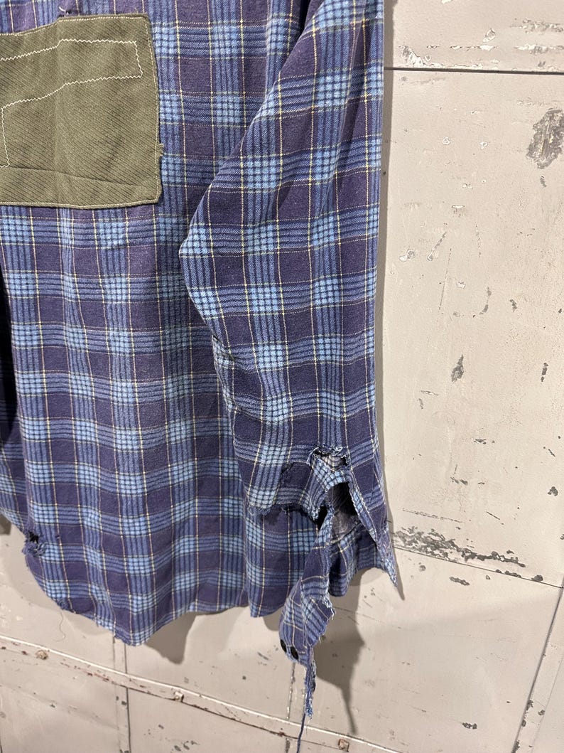 Pu&ograve; includere: Una camicia a maniche lunghe con bottoni e motivo a quadri blu e viola. La camicia presenta una tasca applicata rettangolare color verde oliva sul petto e mostra visibili sfilacciature e danni al tessuto lungo il polsino della manica.