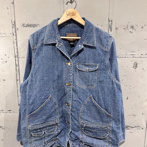 May include: A blue denim jacket with a collared neckline and multiple pockets. The jacket has a button-up front and gold-colored buttons. The jacket is hanging on a wooden hanger with the text "Great Northern Vintage".