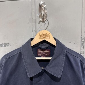 May include: A navy blue Abercrombie jacket with a collar and button closure, hanging on a wooden hanger. The hanger is branded with "Great Northern Vintage". The jacket has a label that reads "Abercrombie 1892".