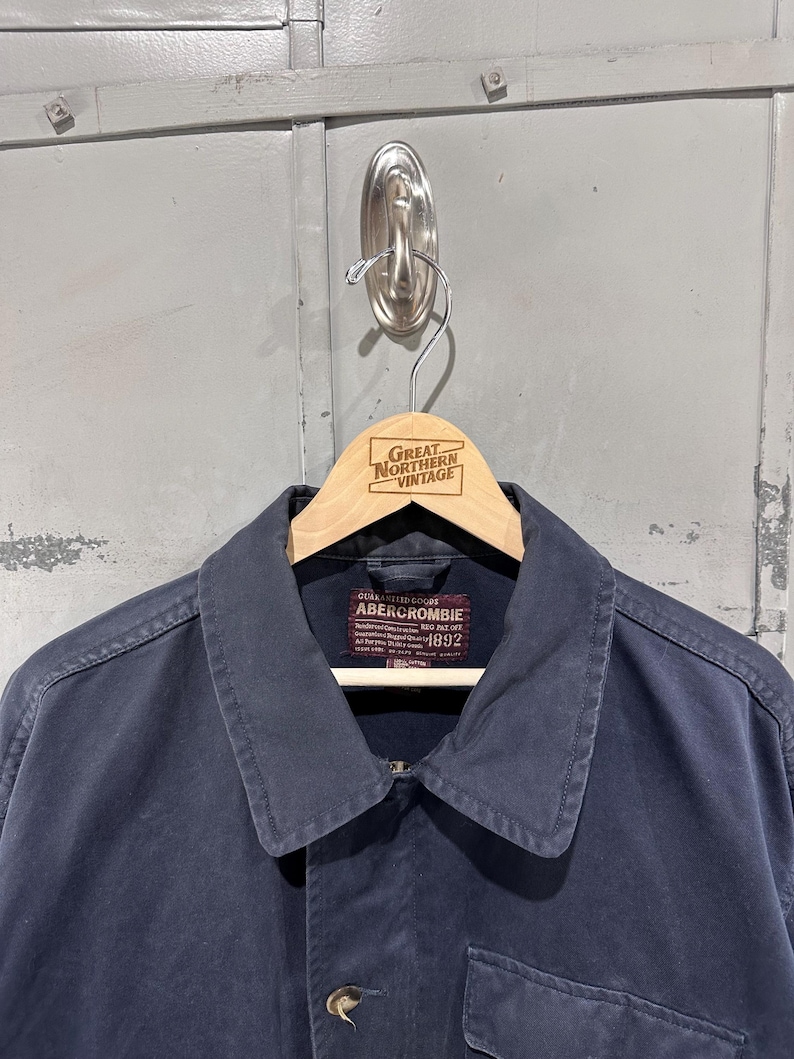 May include: A navy blue Abercrombie jacket with a collar and button closure, hanging on a wooden hanger. The hanger is branded with "Great Northern Vintage". The jacket has a label that reads "Abercrombie 1892".