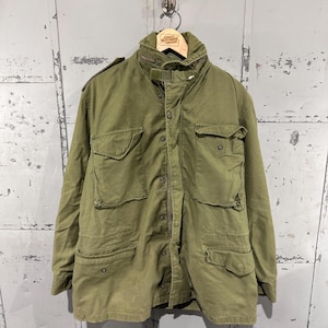 May include: Olive green military-style jacket with multiple front pockets and a button-up closure. The jacket has a high collar and is hanging on a wooden hanger. The jacket is made of a durable fabric and has a vintage aesthetic.