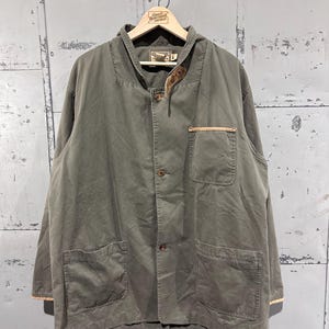 Puede incluir: Chaqueta verde oliva con cuatro bolsillos delanteros y botones. La chaqueta tiene un diseño sin cuello y detalles en cuero marrón en el cuello y los bolsillos. Cuelga de una percha de madera.