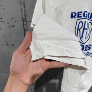 May include: White t-shirt with blue text and a shield graphic. The text reads "REGIONAL" above a shield with "RHS" inside, and "DES" below. The shirt is made of a soft, lightweight fabric.