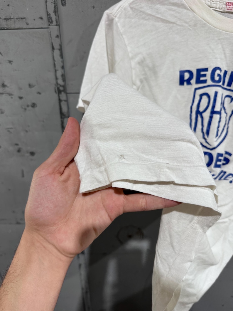 May include: White t-shirt with blue text and a shield graphic. The text reads "REGIONAL" above a shield with "RHS" inside, and "DES" below. The shirt is made of a soft, lightweight fabric.