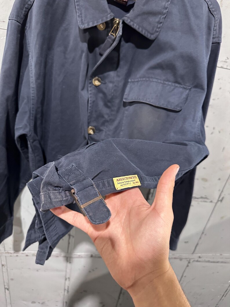 May include: Navy blue jacket with a button-up front, a small chest pocket, and a collar. The jacket has a zipper closure at the neck and a buckle detail on the sleeve. The label reads "Abercrombie".