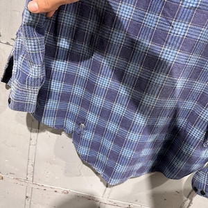 Pu&ograve; includere: Una camicia a maniche lunghe con bottoni caratterizzata da un motivo a quadri blu e azzurro. Il capo presenta un pannello in tessuto verde oliva a contrasto sulla spalla e mostra segni visibili di usura con piccoli fori sfilacciati sull'orlo.