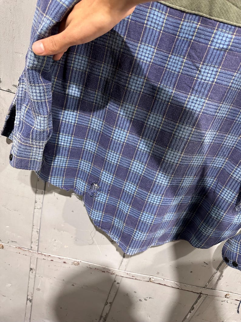 Pu&ograve; includere: Una camicia a maniche lunghe con bottoni caratterizzata da un motivo a quadri blu e azzurro. Il capo presenta un pannello in tessuto verde oliva a contrasto sulla spalla e mostra segni visibili di usura con piccoli fori sfilacciati sull'orlo.