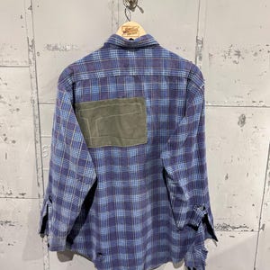 Pu&ograve; includere: Una camicia a maniche lunghe con bottoni in un motivo a quadri blu e viola. Il retro presenta una grande toppa rettangolare in tessuto verde oliva cucita sulla parte superiore. La camicia presenta bordi sfilacciati sui polsini e sull'orlo.