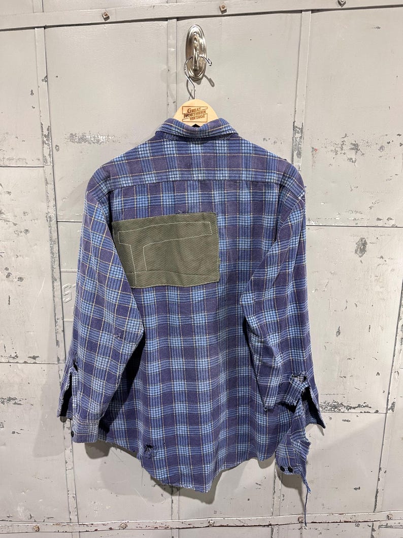 Pu&ograve; includere: Una camicia a maniche lunghe con bottoni in un motivo a quadri blu e viola. Il retro presenta una grande toppa rettangolare in tessuto verde oliva cucita sulla parte superiore. La camicia presenta bordi sfilacciati sui polsini e sull'orlo.