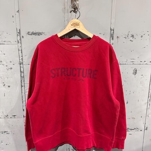 May include: A red crewneck sweatshirt with the word "STRUCTURE" printed in dark navy blue across the chest. The sweatshirt is hanging on a wooden hanger against a gray metal background.