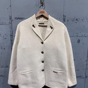 Puede incluir: Chaqueta de lana color crema con cuello con solapas, tres botones oscuros y dos bolsillos delanteros. La chaqueta está colgada en una percha de madera con el texto "Great Northern Vintage".