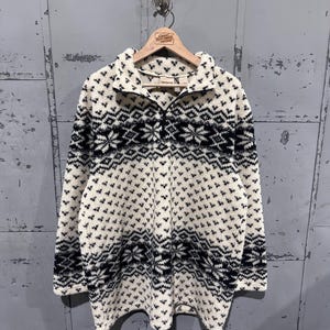 May include: Cream-colored fleece pullover with a black geometric pattern. The long-sleeved top features a half-zip closure and a high collar. The garment is hanging on a wooden hanger against a gray metal background.