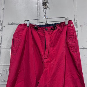 May include: A pair of red cargo trousers with a drawstring waist and multiple pockets. The trousers have a dark blue waistband and a zip fly.