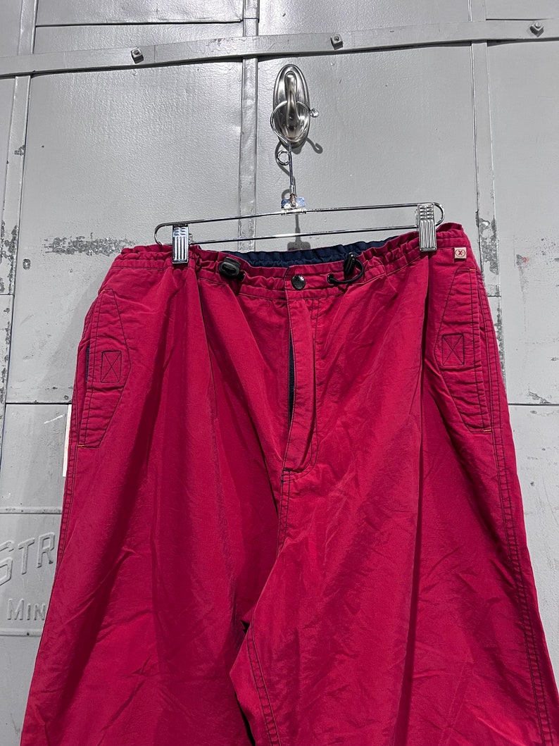 May include: A pair of red cargo trousers with a drawstring waist and multiple pockets. The trousers have a dark blue waistband and a zip fly.