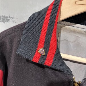 Op de afbeelding: Een close-up van een zwarte varsity-jas met een zwart-rode gestreepte kraag. De jas heeft een gouden rits en een kleine speld met een wapen op de kraag. De jas hangt aan een houten kleerhanger.