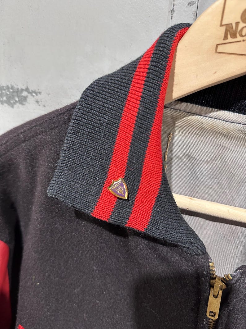 Op de afbeelding: Een close-up van een zwarte varsity-jas met een zwart-rode gestreepte kraag. De jas heeft een gouden rits en een kleine speld met een wapen op de kraag. De jas hangt aan een houten kleerhanger.