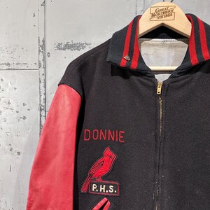 Op de afbeelding: Zwarte en rode vintage varsity jacket met de naam "DONNIE" in het rood geborduurd. Voorzien van een rode kardinaal patch en "P.H.S." patch. De mouwen zijn van rood leer en de kraag heeft rood-zwarte strepen.