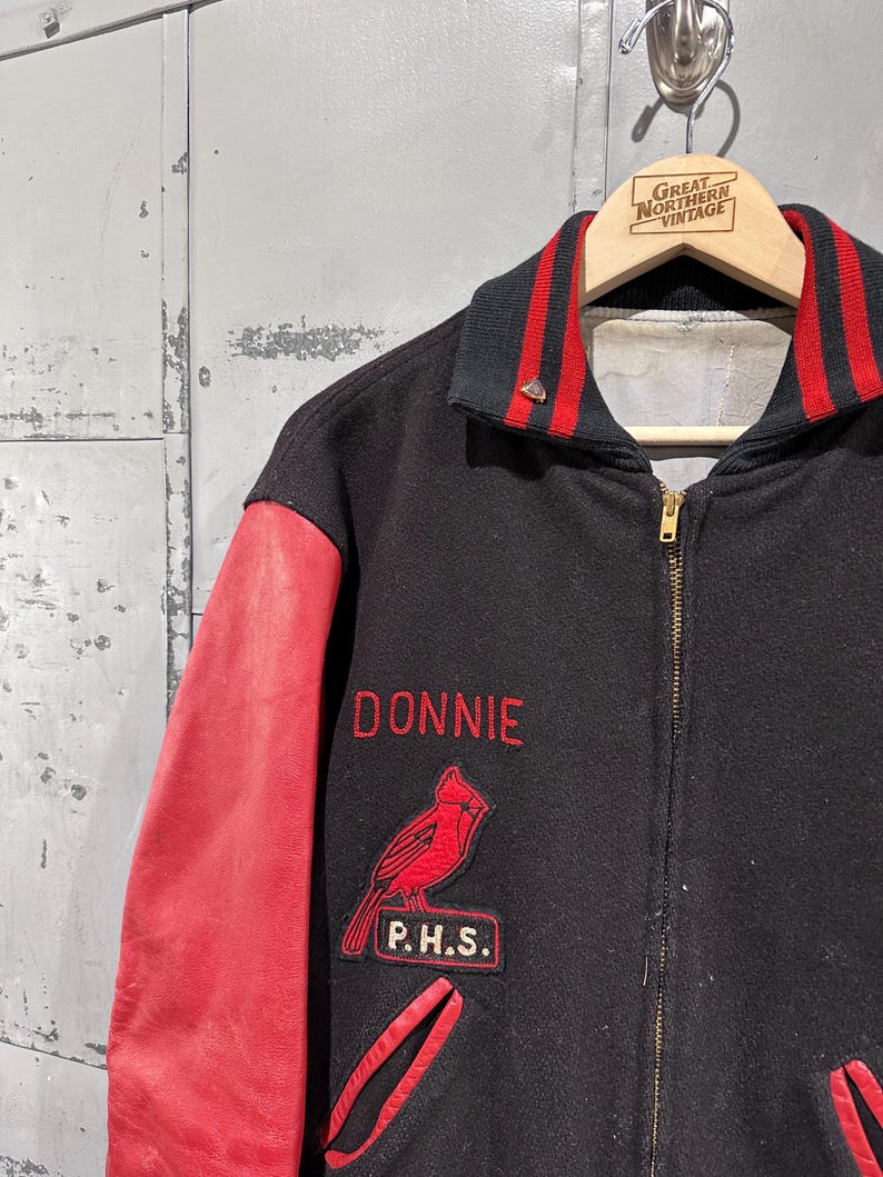 Op de afbeelding: Zwarte en rode vintage varsity jacket met de naam "DONNIE" in het rood geborduurd. Voorzien van een rode kardinaal patch en "P.H.S." patch. De mouwen zijn van rood leer en de kraag heeft rood-zwarte strepen.
