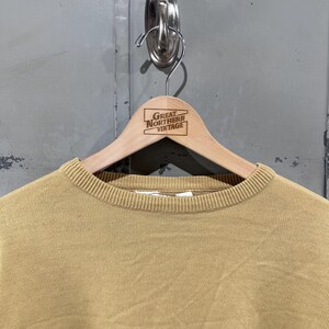 May include: A tan-colored crew neck sweater with the words "Cafe Society" in black script. The sweater is hanging on a wooden hanger with the text "Great Northern Vintage". The background is a gray metal door.