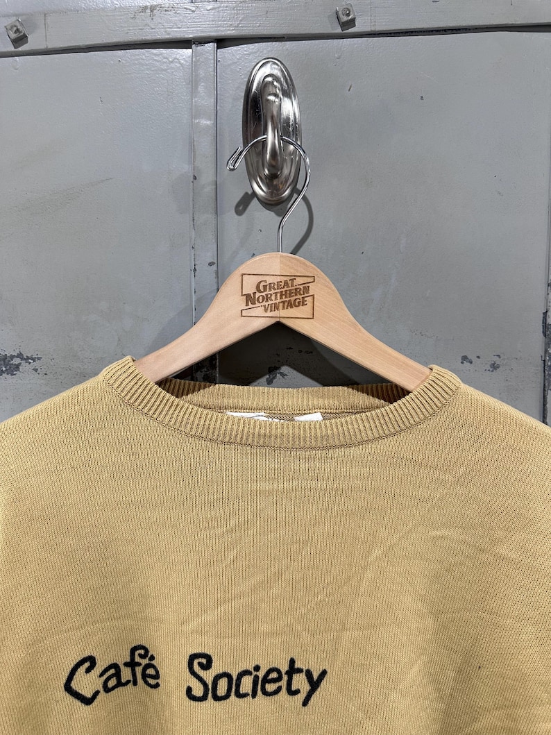 May include: A tan-colored crew neck sweater with the words "Cafe Society" in black script. The sweater is hanging on a wooden hanger with the text "Great Northern Vintage". The background is a gray metal door.