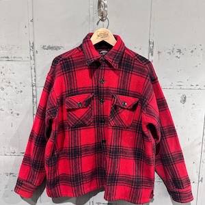 Puede incluir: Una camisa a cuadros rojos y negros con dos bolsillos en el pecho. La camisa está hecha de un material grueso y cálido, perfecto para el clima fresco. La camisa está colgada en una percha de madera.