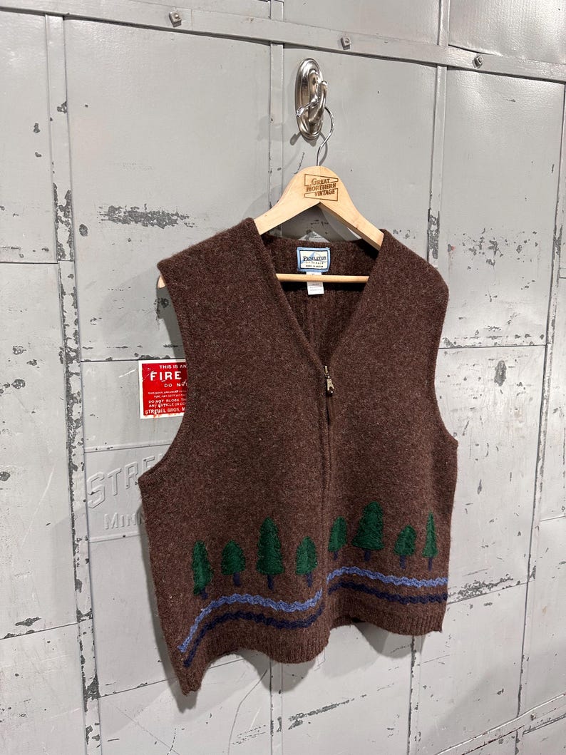 K&ouml;nnte beinhalten: Eine braune Weste aus Wollmischgewebe mit Rei&szlig;verschluss, V-Ausschnitt und einem grafischen Muster am unteren Saum. Das Design zeigt eine Reihe gr&uuml;ner Tannenb&auml;ume &uuml;ber zwei wellenf&ouml;rmigen horizontalen Streifen in Hellblau und Dunkelblau. Pendleton-Etikett.