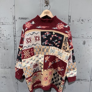 May include: A long-sleeve knit sweater featuring a patchwork design with various patterns including floral motifs, checkered squares, and geometric shapes in a color palette of deep red, cream, black, and tan, finished with a ribbed mock neck collar.