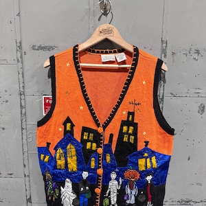 Puede incluir: Chaleco de punto naranja con temática de Halloween, con ribetes y botones negros. Presenta un diseño de paisaje urbano con casas, niños que piden dulces y estrellas. El chaleco está colgado de una percha de madera.